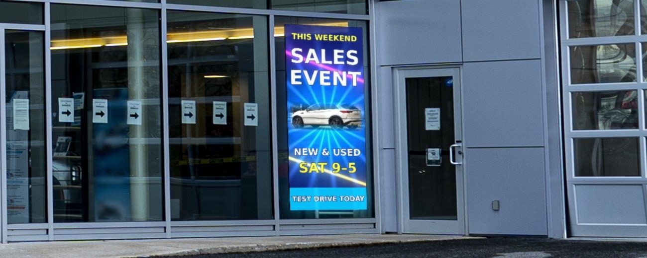 LED screen hung inside, behind a car dealership's storefront glass, facing the street